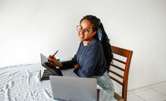 a lady sitting at a table and smiling in from of a laptop and tablet.