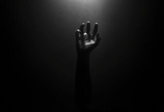 The photo is nearly pitch black, and the faint outline of a hand can be seen reaching upwards towards an unknown light source, as if trying to find hope within the dark