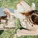 There is a young woman sitting in a field of grass, a mirror in front of her. She is touching her reflection and is looking at herself, as if trying to see inside of herself in regards to who she is.