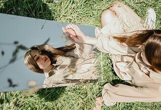 There is a young woman sitting in a field of grass, a mirror in front of her. She is touching her reflection and is looking at herself, as if trying to see inside of herself in regards to who she is.