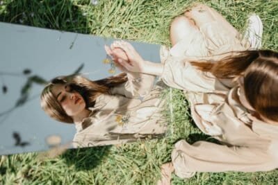 There is a young woman sitting in a field of grass, a mirror in front of her. She is touching her reflection and is looking at herself, as if trying to see inside of herself in regards to who she is.