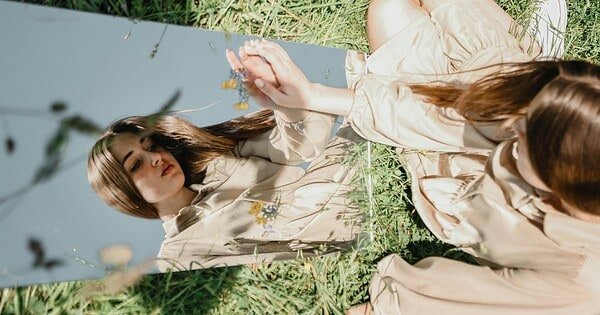 There is a young woman sitting in a field of grass, a mirror in front of her. She is touching her reflection and is looking at herself, as if trying to see inside of herself in regards to who she is.