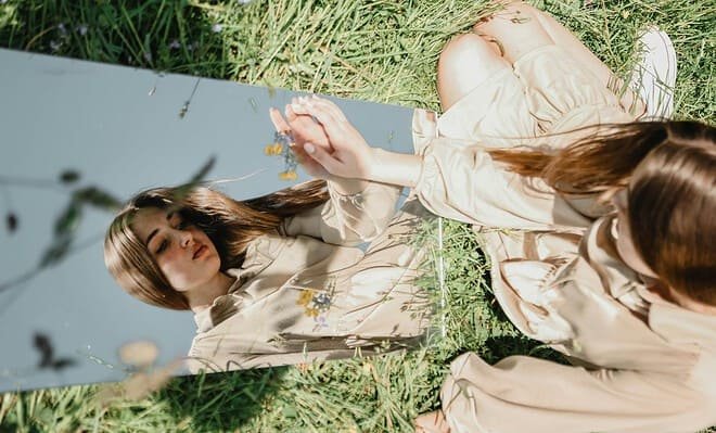 There is a young woman sitting in a field of grass, a mirror in front of her. She is touching her reflection and is looking at herself, as if trying to see inside of herself in regards to who she is.