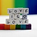 Against a rainbow background, wooden tiles with letters sit on a white table, stacked to spell "love is love" atop rainbow tiles.