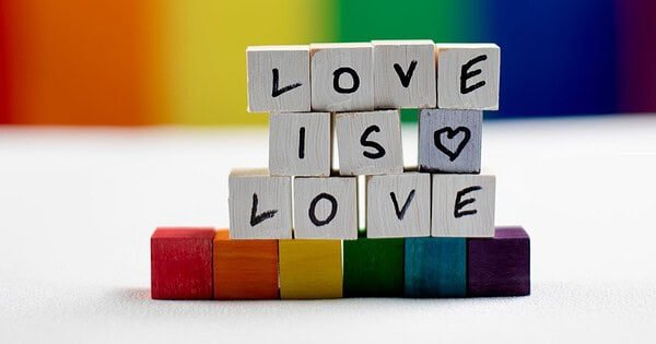 Against a rainbow background, wooden tiles with letters sit on a white table, stacked to spell "love is love" atop rainbow tiles.