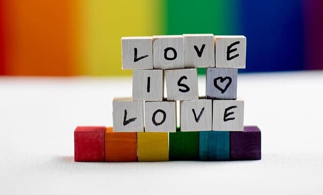 Against a rainbow background, wooden tiles with letters sit on a white table, stacked to spell "love is love" atop rainbow tiles.