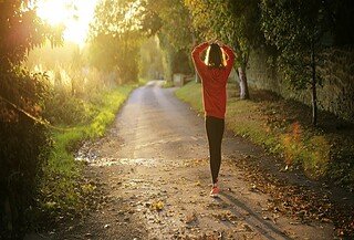 A woman walking in a tree lined forest with a sunlight shining ahead of her.