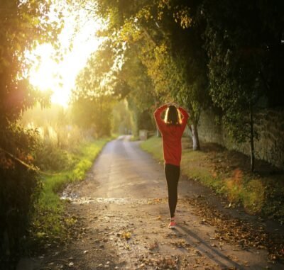 A woman walking in a tree lined forest with a sunlight shining ahead of her.