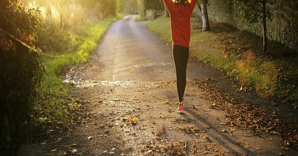 A woman walking in a tree lined forest with a sunlight shining ahead of her.