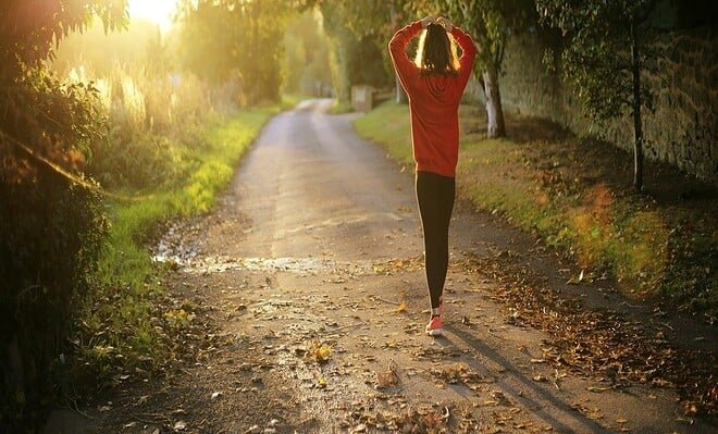 A woman walking in a tree lined forest with a sunlight shining ahead of her.