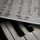 There is a piano shown in the image along with a music sheet that lies slightly atop the piano, faint shadows shown underneath the paper as well.