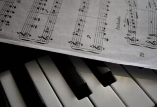 There is a piano shown in the image along with a music sheet that lies slightly atop the piano, faint shadows shown underneath the paper as well.