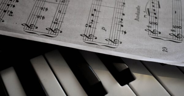 There is a piano shown in the image along with a music sheet that lies slightly atop the piano, faint shadows shown underneath the paper as well.