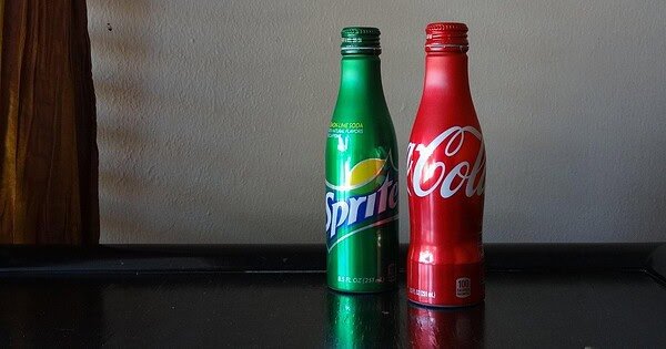 A red Coke bottle and green Sprite bottle sit against a white wall