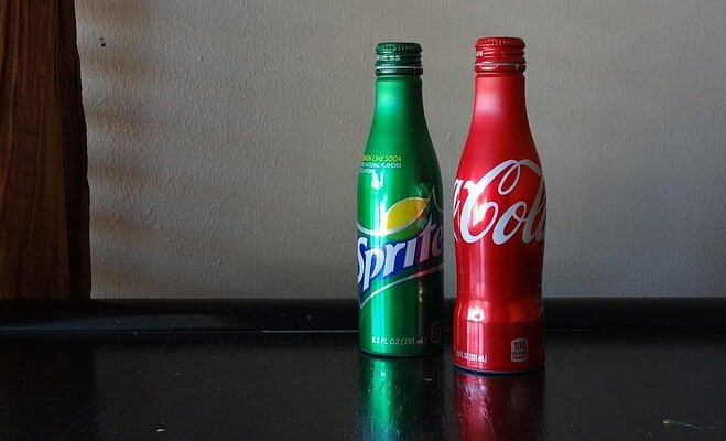 A red Coke bottle and green Sprite bottle sit against a white wall