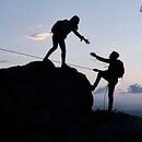 Two silhouettes reach the top of a mountain, backed by the soft light of the setting sun