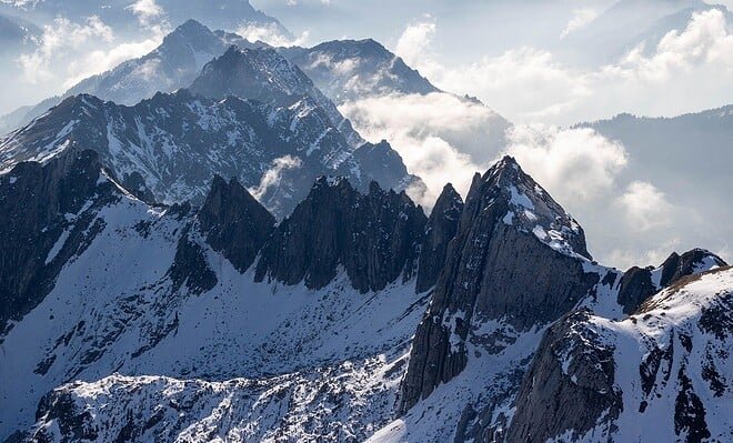 Snow covered mountain peeks along a blue sky horizon with low hanging clouds