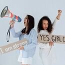 There is a light background and two women standing back to back, holding signs that say "I am strong" and "Yes Girl Can" as they are chanting about the power they hold as women. One is also holding a megaphone as they are chanting.