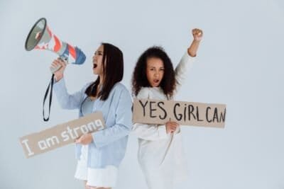 There is a light background and two women standing back to back, holding signs that say "I am strong" and "Yes Girl Can" as they are chanting about the power they hold as women. One is also holding a megaphone as they are chanting.