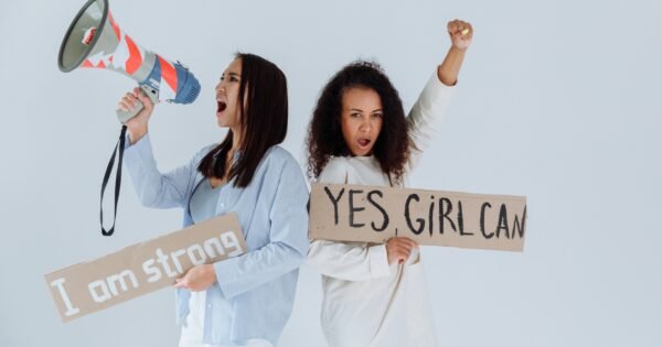 There is a light background and two women standing back to back, holding signs that say "I am strong" and "Yes Girl Can" as they are chanting about the power they hold as women. One is also holding a megaphone as they are chanting.