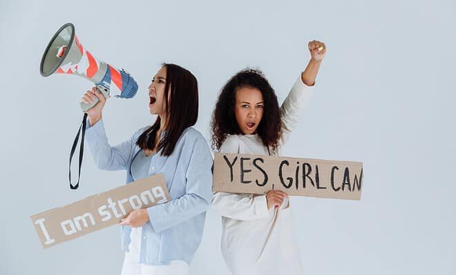 There is a light background and two women standing back to back, holding signs that say "I am strong" and "Yes Girl Can" as they are chanting about the power they hold as women. One is also holding a megaphone as they are chanting.