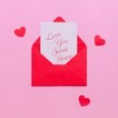 A pink background is set with small red hearts around a small envelope that is opened with a note peaking out that reads "Love you sweet heart"