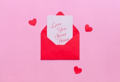 A pink background is set with small red hearts around a small envelope that is opened with a note peaking out that reads "Love you sweet heart"
