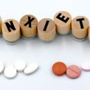 There's a white background with wooden pieces that have letters on them, spelling out the word Anxiety. Below the word is two groups of pills, likely representing anti anxiety medication.