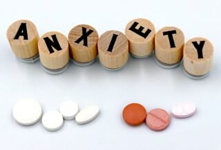There's a white background with wooden pieces that have letters on them, spelling out the word Anxiety. Below the word is two groups of pills, likely representing anti anxiety medication.