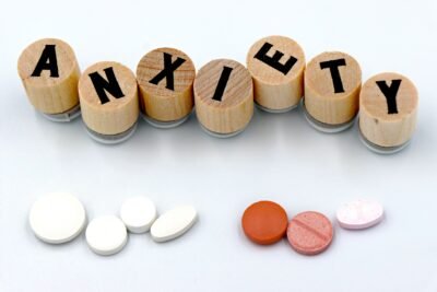 There's a white background with wooden pieces that have letters on them, spelling out the word Anxiety. Below the word is two groups of pills, likely representing anti anxiety medication.