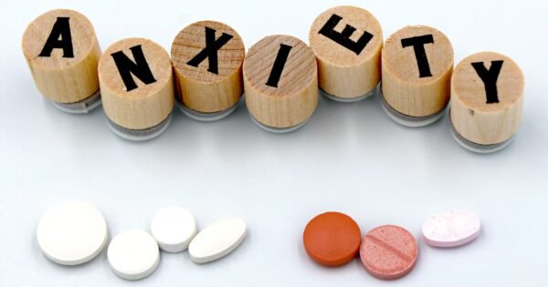 There's a white background with wooden pieces that have letters on them, spelling out the word Anxiety. Below the word is two groups of pills, likely representing anti anxiety medication.