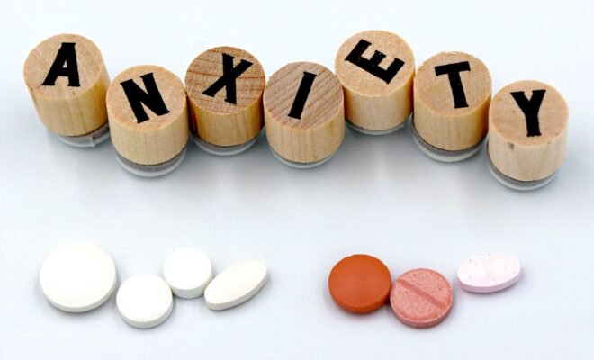 There's a white background with wooden pieces that have letters on them, spelling out the word Anxiety. Below the word is two groups of pills, likely representing anti anxiety medication.