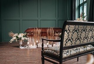 There is a room with a closed casket, surrounded by plants and with a bench sitting in front of it, as if showing a funeral service being prepared for someone who has passed away