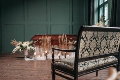 There is a room with a closed casket, surrounded by plants and with a bench sitting in front of it, as if showing a funeral service being prepared for someone who has passed away