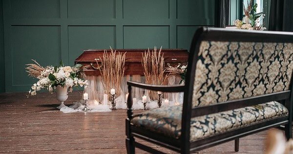 There is a room with a closed casket, surrounded by plants and with a bench sitting in front of it, as if showing a funeral service being prepared for someone who has passed away