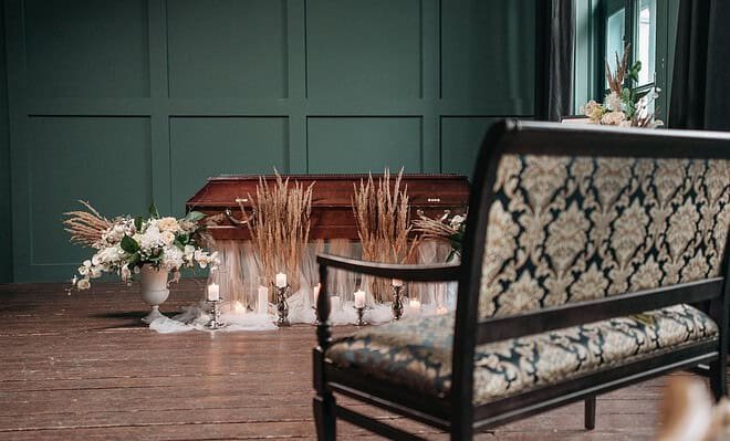There is a room with a closed casket, surrounded by plants and with a bench sitting in front of it, as if showing a funeral service being prepared for someone who has passed away