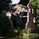 a ruined brick house sits on the bank of a river surrounded by green trees.