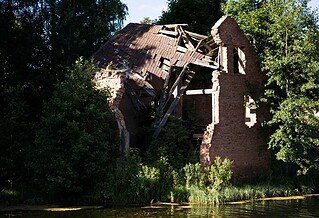 a ruined brick house sits on the bank of a river surrounded by green trees.
