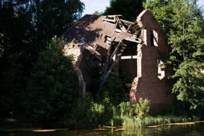 a ruined brick house sits on the bank of a river surrounded by green trees.
