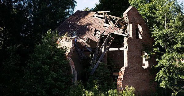 a ruined brick house sits on the bank of a river surrounded by green trees.