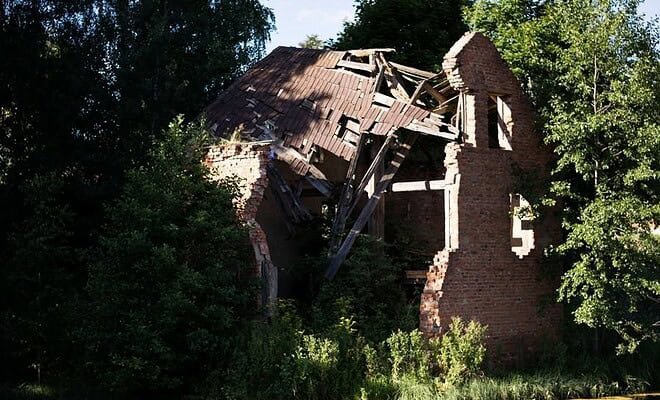 a ruined brick house sits on the bank of a river surrounded by green trees.