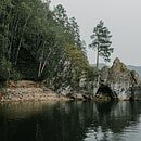 Tall pine trees give way to a rocky shoreline which meets lake, whose calm waters reflect the gray sky.