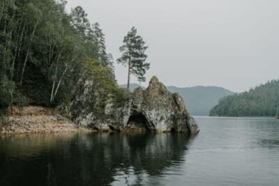 Tall pine trees give way to a rocky shoreline which meets lake, whose calm waters reflect the gray sky.